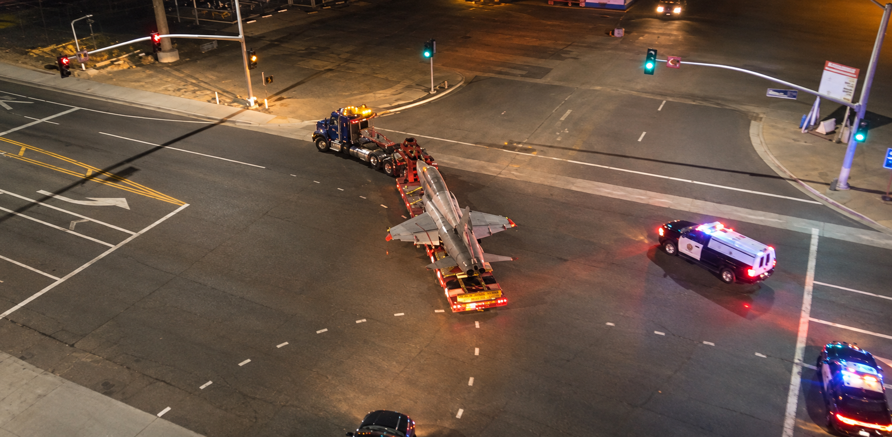 Nighttime overhead view of the aircraft on a transport trailer turning through an intersection with escort vehicles